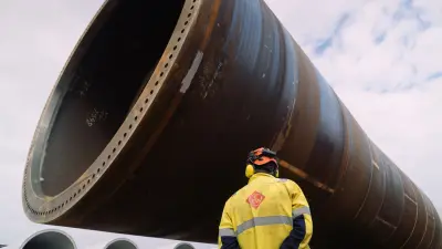 A worker in the harbor looks up at a massive, horizontal monopile foundation.