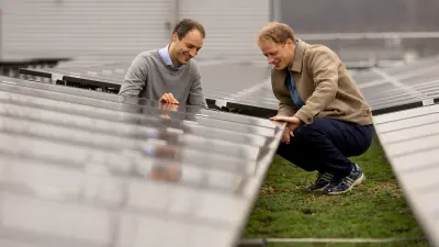 Two men are smiling as they inspect a row of solar panels on a green roof.